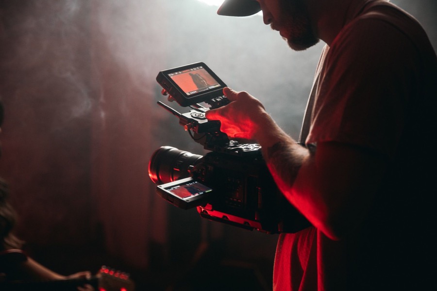 Mann mit Baseballkappe bedient eine Videokamera mit externem Monitor in rauchiger, rot beleuchteter Umgebung.