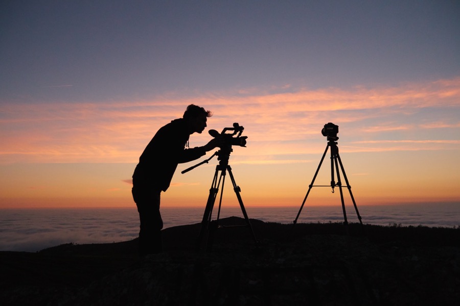 Silhouette eines Kameramanns bei Sonnenaufgang mit zwei Kameras auf Stativen, über Wolken.