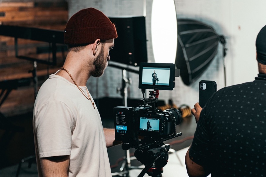 Ein Mann mit roter Mütze und weißem T-Shirt bedient eine Videokamera mit externem Monitor; ein zweiter Mann filmt mit einem Smartphone.