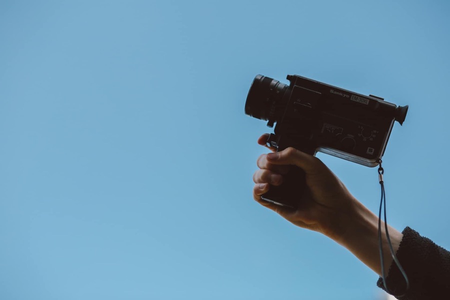 Eine Hand hält eine schwarze Vintage-Videokamera mit einem Objektiv vor einem klaren blauen Himmel.